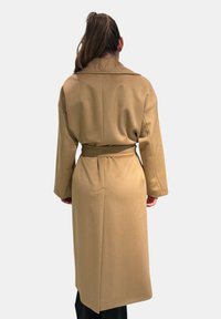 Cappotto lungo beige con cintura, design con colletto, tessuto leggermente strutturato, maniche lunghe e spacco posteriore per facilitare i movimenti.