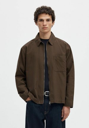 Jeune homme aux cheveux bouclés portant une veste zippée à carreaux marron sur un t-shirt noir et un jean bleu foncé, debout devant un fond blanc uni.
