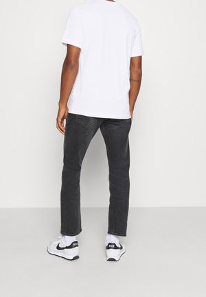 Mann in weißem T-Shirt, dunklen grauen Jeans und weißen Nike-Sneakern, der vor einem einfachen hellen Hintergrund steht und von der Kamera wegschaut.