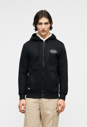 Giovane uomo che indossa una felpa nera con zip e logo "Von Dutch" e pantaloni cargo beige, in piedi contro uno sfondo chiaro e semplice.