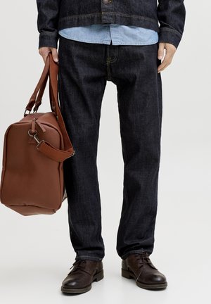 Homme portant un jean foncé, des chaussures en cuir marron et une chemise en denim sous une veste en denim, tenant un sac de voyage en cuir marron.
