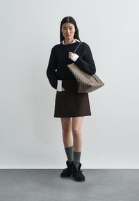 Black knit sweater with white collar, brown suede skirt, gray socks, and black shoes. Holding a patterned tote bag. Minimalist background.