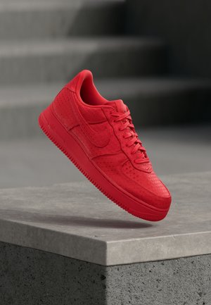 Baskets Nike Air Force 1 rouges avec empeigne texturée et lacets, positionnées en l'air au-dessus d'un bloc de béton sur un fond d'escaliers floutés.