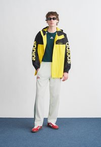 Κίτρινο και μαύρο αθλητικό μπουφάν τύπου windbreaker με το λογότυπο "adidas", πράσινο T-shirt, λευκό παντελόνι με ρίγες στο πλάι και κόκκινα αθλητικά παπούτσια.