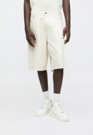 Mand iført knælange off-white shorts, hvide sokker og hvide sneakers foran en ensfarvet lys baggrund.