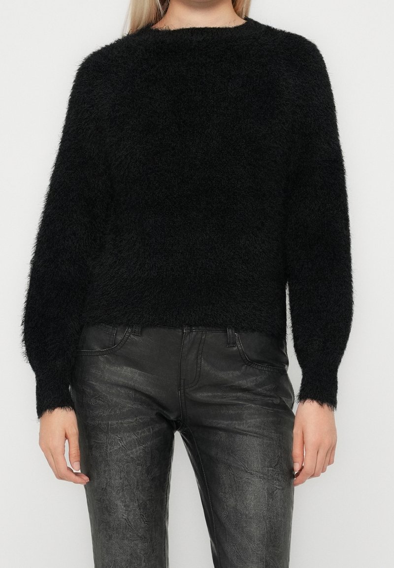 Personne portant un pull noir duveteux à manches longues et un pantalon en cuir noir texturé, debout devant un fond clair uni.