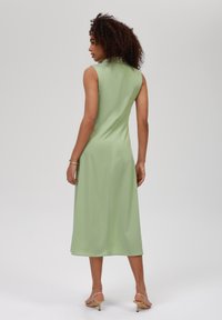 Abito verde senza maniche con colletto alto e lunghezza midi, caratterizzato da una texture liscia e da un sottile dettaglio sulle cuciture laterali.
