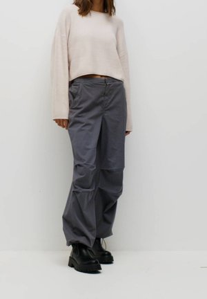 Person iført beige langærmet cropped sweater, grå løstsiddende bukser og sorte chunky støvler mod en ensfarvet hvid baggrund.