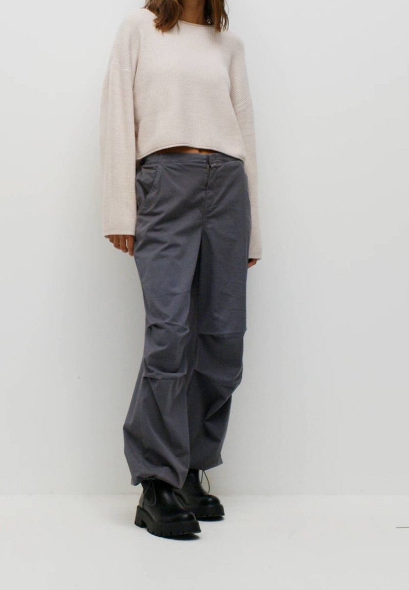 Personne portant un pull court beige à manches longues, un pantalon ample gris et des bottes noires épaisses sur un fond blanc uni.