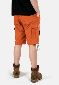 Oranger Cargoshorts mit mehreren Taschen und Kordelzugdetails, aus einem strukturierten Stoff, kombiniert mit braunen Stiefeln. Ansicht von hinten.