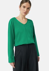 Groene long-sleeve V-hals top van gladde stof, met een subtiele naad aan de voorkant, gecombineerd met zwarte op maat gemaakte broeken.