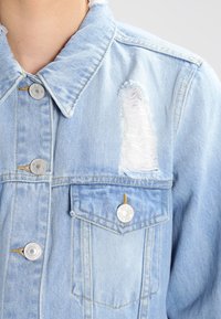 Ljusblå denimjacka med silverfärgade knappar, bröstficka och en stor sliten reva ovanför fickan på högra sidan.