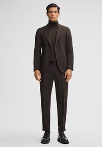 Dunkelbrauner Anzug mit taillierter Jacke und Hose, strukturiertem Stoff und passendem Rollkragenpullover. Schwarze Schnürschuhe runden das Outfit ab.