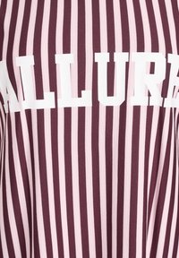 Maroonowe i różowe pasiasty materiał z białymi literami blokowymi piszącymi "ALLURE". Gładka tekstura, pionowe paski równomiernie rozmieszczone.
