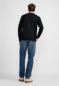 Maglione a righe scure con maniche lunghe, abbinato a jeans blu e sneakers chiare, visto da dietro su uno sfondo semplice.