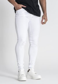 Jeans ajustados blancos con detalles desgatados, que cuentan con un diseño ceñido y una textura suave, combinados con zapatillas blancas.