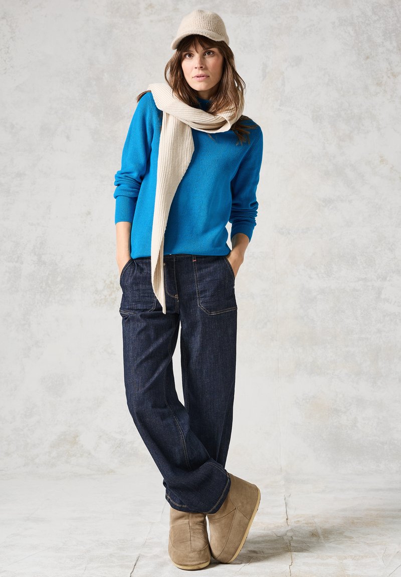 Helle blaue Strickjacke, cremefarbener Schal drapiert, dunkelblaue weit geschnittene Jeans und beige Stiefel. Eine Mütze aus passendem cremefarbenen Material vervollständigt das Outfit.
