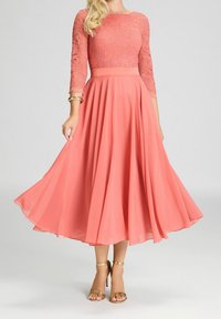 Robe corail avec un corsage en dentelle, des manches trois-quarts et une jupe fluide. Tissu lisse avec une taille marquée, assortie à des sandales dorées.