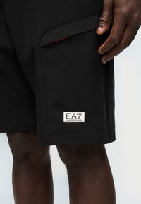 Μαύρα σορτς με φερμουάρ σε τσέπη, φορεμένα από ένα άτομο, με λευκό λογότυπο EA7 Emporio Armani κοντά στο τελείωμα στο αριστερό πόδι.