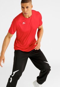 Rode sportieve t-shirt met een ronde hals en logo. Gecombineerd met zwarte broeken met witte zijaccenten. Glad materiaal, sportieve pasvorm.