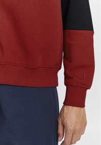 Rød og svart hettegenser med ribbede mansjetter og fald, med en glatt tekstur og kontrasterende sleeve-design. Synlig hånd som hviler på stoffet.