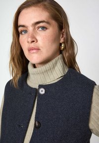 Gilet di lana blu navy con scollatura rotonda, dotato di due bottoni e un dolcevita testurizzato di colore crema sottostante. Design semplice e pulito.