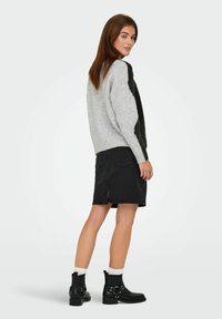 Femme debout, portant un pull gris et noir, une jupe en denim noire avec une fente arrière, des chaussettes blanches et des bottines noires contre un fond uni.