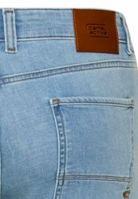 Jasnoniebieska tylna kieszeń jeansów z denimu z brązową skórzaną naszywką z logo "come.active" na pasku talii.