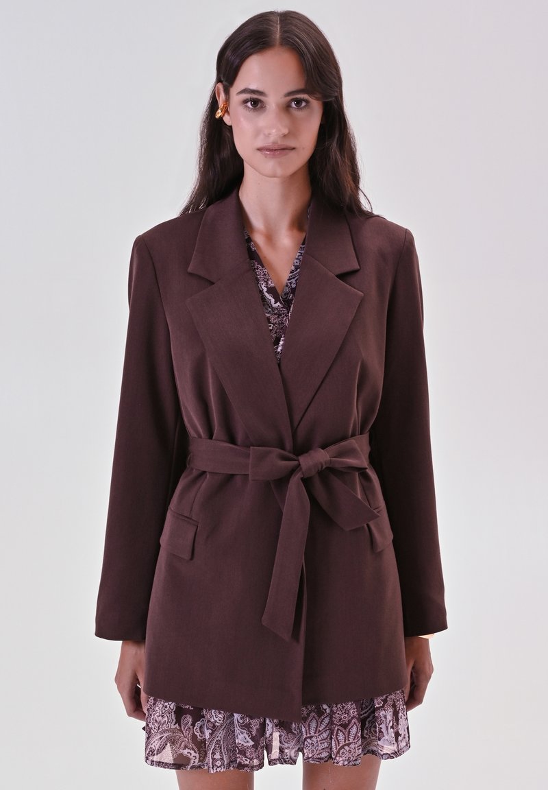 Bruine blazer met riem, oversized revers en twee voorzakken, gedragen over een lichter patroonjurk met een gerafeld zoom.