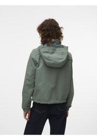 Personne aux cheveux bouclés portant une veste à capuche verte et un pantalon sombre, vue de dos sur un fond uni.