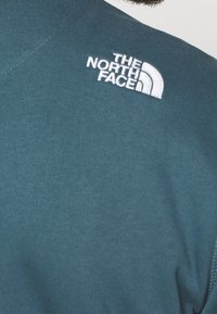 Sweatshirt teal à manches courtes, avec un logo brodé blanc de The North Face sur l'épaule gauche. Texture lisse.
