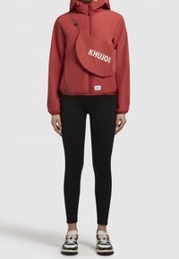 Red lightweight jacket with hood, front zipper, and embroidered "KHUJO 2004" shoulder bag. Black leggings and sneakers complete the outfit.