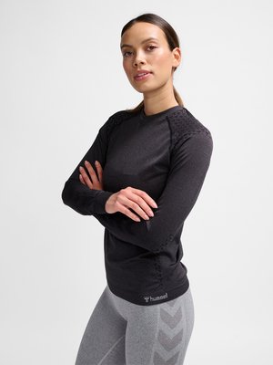 Jonge vrouw met gekruiste armen, die een zwarte lange mouwen sporttop en grijze legging met chevronpatronen draagt tegen een witte achtergrond.