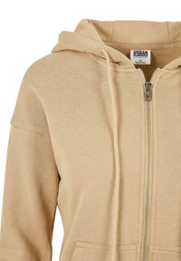 Beige zip-up hette laget av mykt stoff, med hette med snor, frontlommer og sølvglidelås. Teksturert, uformell design.
