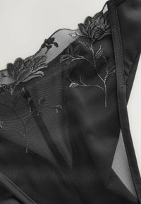 Schwarzer Spitzen-Thong mit floralem Stickerei, durchsichtigem Mesh-Obermaterial und glattem Stoff. Detaillierte Nähte entlang der Kanten und florale Akzente.