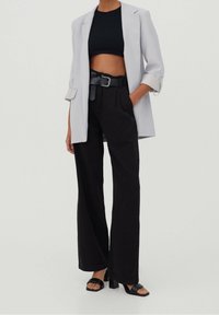 Personne portant un blazer gris clair, un crop top noir, un pantalon noir taille haute avec ceinture, et des sandales à talons noires, debout devant un fond uni.