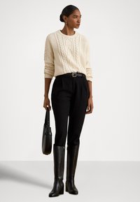 Krémový sveter s vrkočovým vzorom, čierne nohavice s vysokým pásom a opaskom, a čierne kožené čižmy nad kolená. Drží okrúhlu čiernu kabelku.