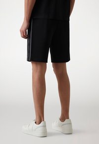 Czarne sportowe szorty z bocznymi paskami, wykonane z miękkiego materiału. Noszone z białymi sneakersami. Widok z tyłu, ukazujący długość i dopasowanie.