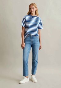 Jovem mulher a usar uma t-shirt riscada azul e branca, jeans claros, ténis brancos e um colar de prata, de pé contra um fundo simples.