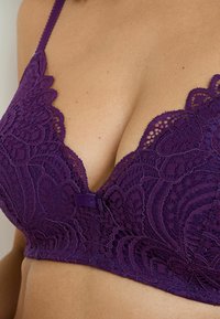 Brassiere in pizzo viola con scollatura profonda, motivi floreali, bordi ondulati e un'accentuata fiocco centrale.