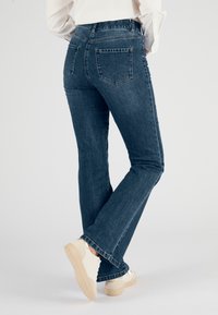 Jeans de mezclilla azul oscuro con un corte relajado, que presentan bolsillos traseros, detalles de costura y un diseño de pierna acampanada. Llevados con zapatillas de color claro.