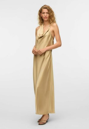 Kobieta o kręconych blond włosach, ubrana w złotą satynową suknię maxi na szyi typu halter oraz buty na otwarte palce, stojąca na tle jednolitego, jasnego tła.