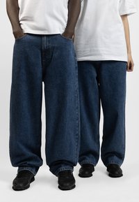 Mörkblå vida denimjeans med en avslappnad passform, med framfickor och bärs med svarta sneakers. Texturen verkar jämn med minimal blekning.