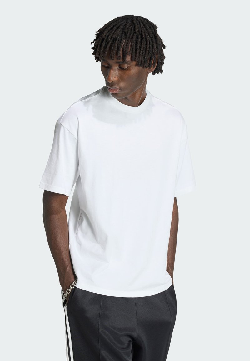 Mand med korte dreadlocks iført en simpel hvid T-shirt med korte ærmer og sorte bukser, kigger ned med hænderne i lommerne.