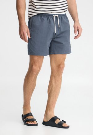 Uomo che indossa pantaloncini blu navy con coulisse, maglietta a righe e sandali neri non abbinati in piedi su sfondo bianco.