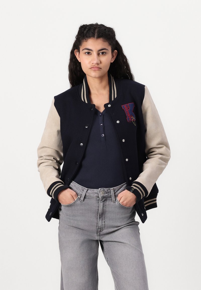 Marineblaues Varsity-Jackett mit beigen Ärmeln, gerippten Bündchen und Kragen. Mit einem roten "K"-Logo und metallenen Druckknöpfen. Tragend über einem schwarzen Oberteil.
