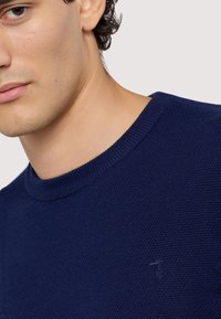 Marinblå stickad tröja med en texturerad yta, med en rund halsringning och subtil logo-detalj på bröstet.