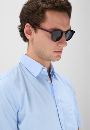 Giovane uomo con capelli scuri che indossa occhiali da sole neri e una camicia azzurro chiaro con bottoni su uno sfondo chiaro e semplice.