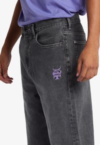 Čierne džínsy z denimu s voľným strihom. Obsahujú fialovo vyšívané logo na prednej kapse a kontrastné švy. Hladká textura, dĺžka po členky.