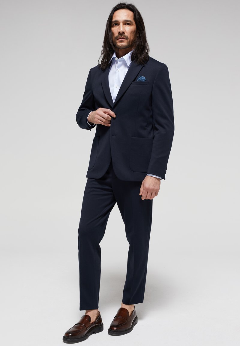 Traje azul marino a medida con solapas de muesca, dos bolsillos delanteros y una camisa blanca. Presenta mocasines marrones y un pañuelo de bolsillo azul.